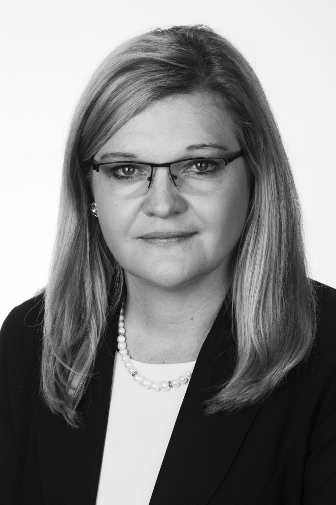 Black and white portrait of a woman with light hair, wearing glasses, a dark blazer, a light top, and a beaded necklace, looking directly at the camera with a neutral expression.